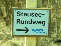 ein Wegweiser mit der Aufschrift "Stausee-Rundweg". 3 blaue Wellenlinien kennzeichnen den Weg.